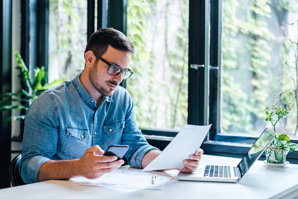 Młody mężczyzna w okularach siedzi przy biurku, na którym leży otwarty laptop. Mężczyzna w jednym ręku trzyma smartfon, a w drugim dokument, który czyta